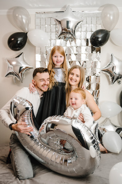 爸爸妈妈恭贺女儿，儿子生日近拱门。妈妈，爸爸抱着孩子。幸福的家庭庆祝6年生日聚会。照片墙装饰白色、黑色、银色气球。图片下载