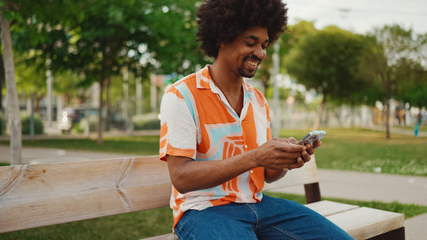 一个穿着衬衫的非洲裔美国年轻人坐在公园长椅上玩智能手机的特写。一个微笑的非洲裔美国人看手机上的照片视频的特写。生活方式的概念图片下载