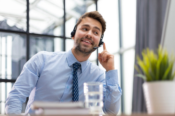 开朗的年轻支持电话男接线员戴着耳机，在工作场所与同事一起使用电脑，帮助服务和客户咨询呼叫中心的概念。图片下载