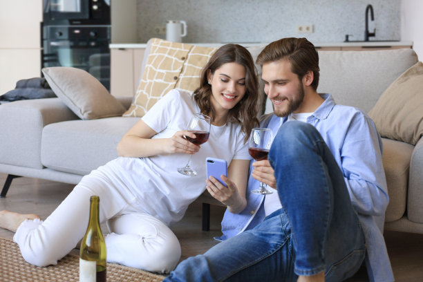 幸福的夫妇坐在客厅的地板上，喝着红酒。年轻的丈夫和妻子微笑着在家里休息，享受家庭周末的浪漫约会图片下载