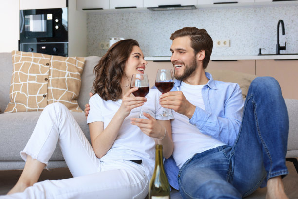 幸福的夫妇坐在客厅的地板上，喝着红酒。年轻的丈夫和妻子微笑着在家里休息，享受家庭周末的浪漫约会图片下载