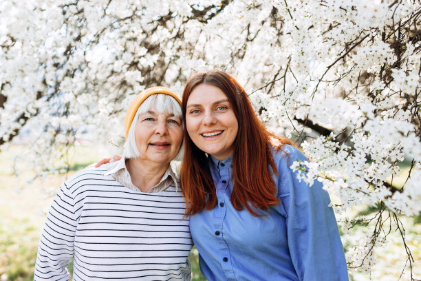 幸福的爱年长成熟的母亲和女儿笑拥抱，有爱心的年轻女子拥抱老年中年妈妈一起度过的时光。树上白色美丽的花朵在春天盛开图片下载