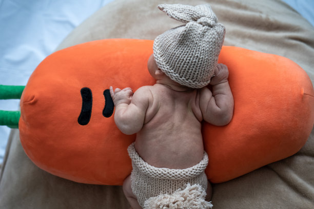 可爱的新生婴儿穿着米色针织兔服躺在橙色毛绒枕头上，婴儿在胡萝卜玩具上爬行，学习走路图片下载