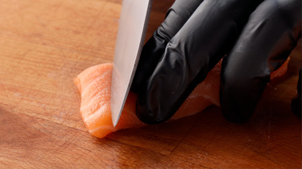 一个戴着黑色乳胶手套的厨师用一把锋利的刀切着一条红色的鱼。切板上的红鲑鱼片。图片下载