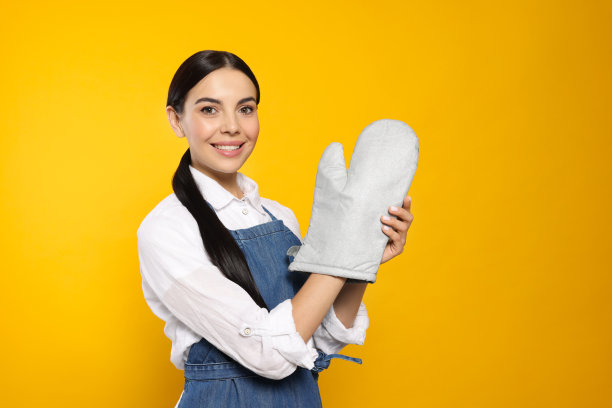 穿着蓝色牛仔裤，围裙，戴着烤箱手套的黄色背景的年轻女子图片下载