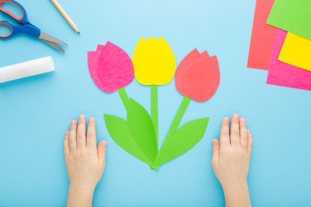 小孩子的手和五颜六色的郁金香花与绿叶从纸上浅蓝色的桌子背景。柔和的颜色。视角镜头。特写镜头。制作装饰元素。自顶向下视图。图片下载