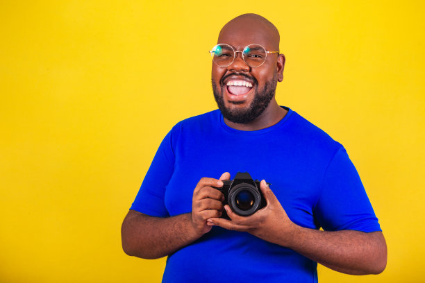 英俊的非洲裔巴西人，戴着眼镜，黄底蓝衬衫。拿着相机，摄影师，微笑，创意。数码单反相机图片下载