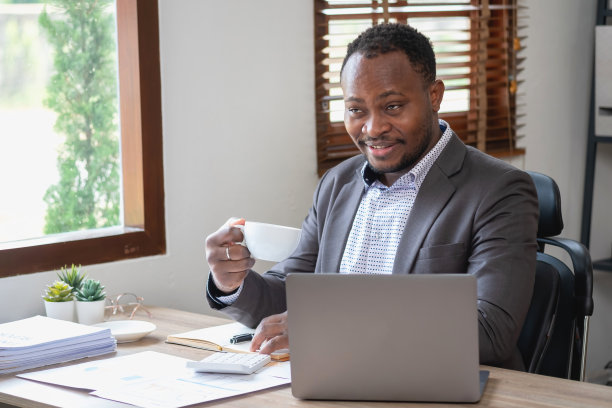 一位身穿黑色西装的非洲裔美国商人，在办公室里用笔记本电脑查看公司工作后，端着一杯咖啡放松一下。图片下载