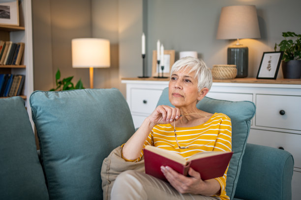 一位退休妇女舒适地坐在公寓的沙发上，全神贯注地看她最喜欢的书图片下载
