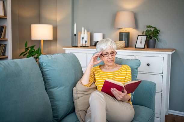 一位退休妇女舒适地坐在公寓的沙发上，全神贯注地看她最喜欢的书图片下载