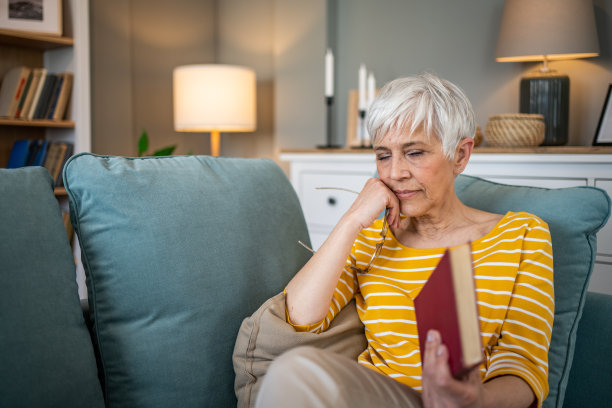 一位退休妇女舒适地坐在公寓的沙发上，全神贯注地看她最喜欢的书图片下载