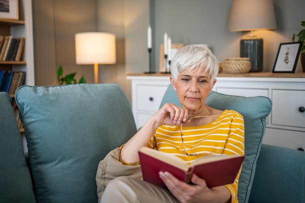 一位退休妇女舒适地坐在公寓的沙发上，全神贯注地看她最喜欢的书图片下载