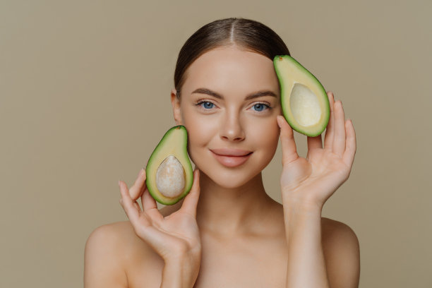 照片中，赤裸上身的女模特在镜头前表情温和，近脸有一半的牛油果，在米色背景下进行了美容处理，皮肤健康，保养得很好。图片下载