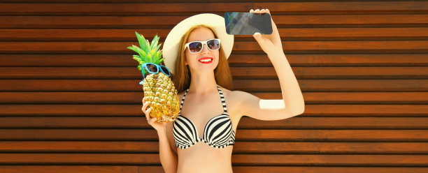 夏日肖像，快乐的女人带着草帽、太阳镜和胸罩，拿着菠萝用智能手机自拍图片下载