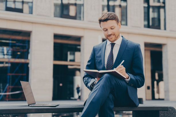 穿着蓝色西装的微笑年轻人在户外时在笔记本上做笔记图片下载