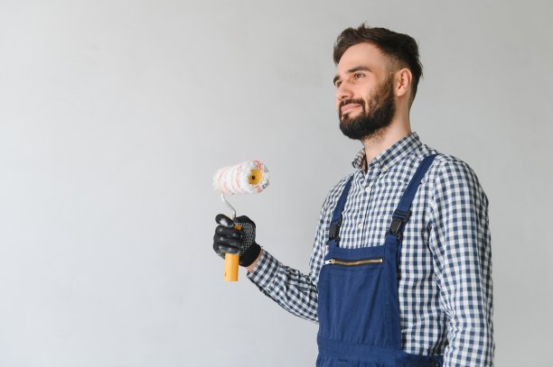 穿着蓝色制服的年轻专业工人微笑着站在新公寓的油漆滚筒上，在灰色的墙壁背景上进行修复，复制空间。图片下载