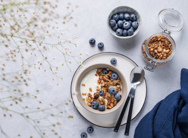 健康早餐的概念是酸奶，格兰诺拉麦片和新鲜蓝莓在蓝色背景上配上鲜花。自制饮食和蛋白质食品。图片下载