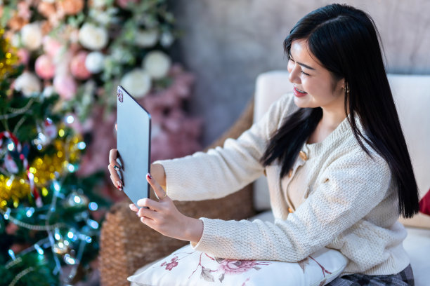 肖像可爱美丽的积极的微笑年轻的亚洲妇女使用平板电脑视频通话或自拍在家里在客厅在室内装饰圣诞节期间，圣诞和玫瑰背景图片下载