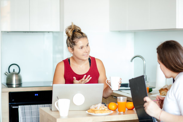 远程工作。两个漂亮的女人拿着笔记本电脑面对面坐着，中间是橙汁，还有新鲜的糕点。图片下载