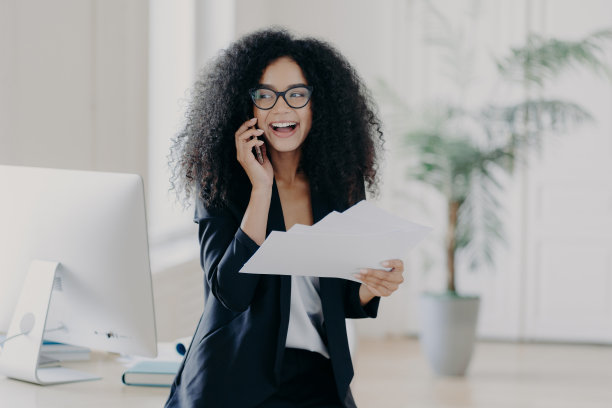 愉快的卷发女董事穿着正装，拿着文件，用手机打电话，站在工作场所附近用电脑，分享创业心得，做业务报告图片下载