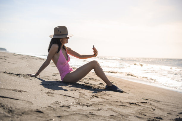 一名年轻的成年女子坐在沙滩上逆光用智能手机打视频电话图片下载
