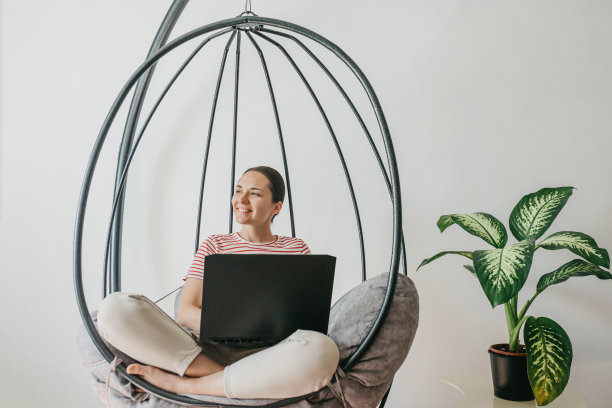 一个年轻漂亮的欧洲女人，坐在家里的吊椅上，用笔记本电脑在家里远程工作。图片下载
