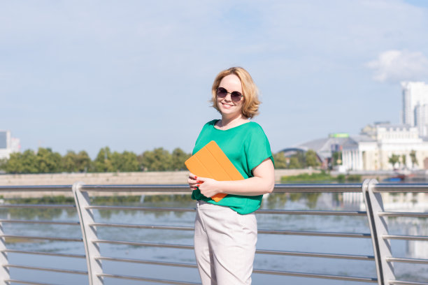 一个面带微笑的博主女孩戴着墨镜，手里拿着笔记本或平板电脑站在河堤上。图片下载
