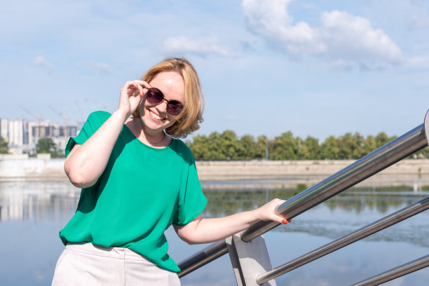 一个戴着墨镜、穿着绿色衬衫、微笑着的快乐女孩站在河岸上的肖像。图片下载