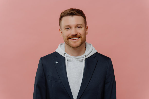 室内拍摄帅气的白人男子，穿着连帽衫和黑色夹克，笑容满面，表情开朗平静，在粉红色的背景下愉快地交谈。人类面部表情图片下载