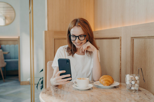 年轻乐观的红发女人戴着耳机坐在咖啡馆里用智能手机上网聊天图片下载