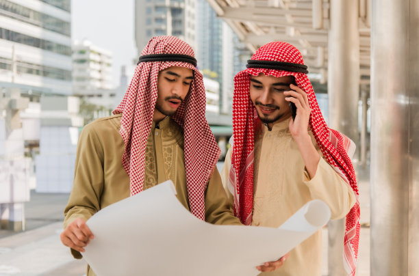 图为两名阿拉伯商人手持蓝图，用智能手机在城市建筑工地开会交谈。图片下载