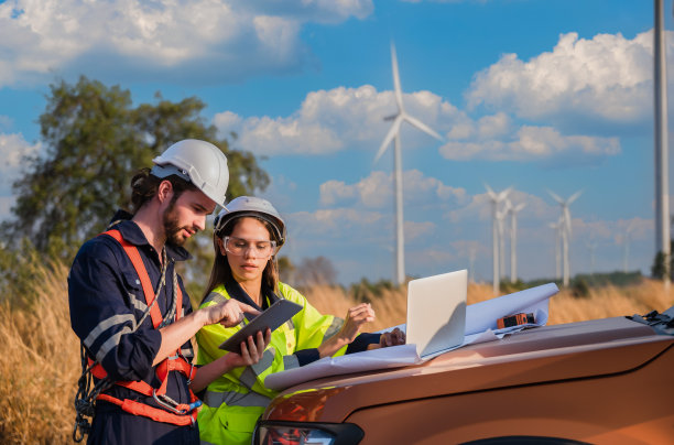 两名专业维护工程师穿着安全工作服，使用笔记本电脑和平板电脑检查风车建设农场的风力涡轮机。图片下载