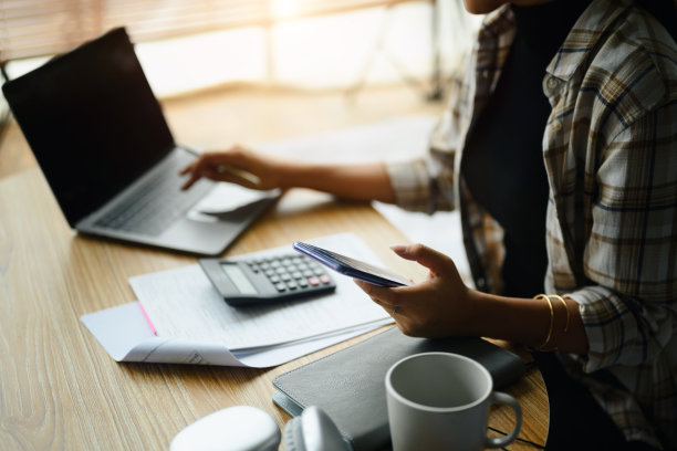 一名年轻女子使用笔记本电脑，通过智能手机在网上银行应用程序上进行支付。图片下载