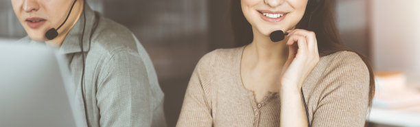 在阳光明媚的现代办公室里，两个戴着耳机微笑着的人坐在办公桌前与客户交谈图片下载