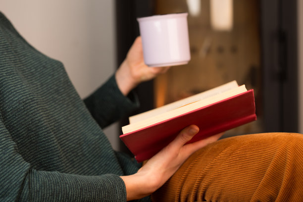 一个年轻的女人正坐在壁炉旁的椅子上看书，手里端着一杯茶图片下载