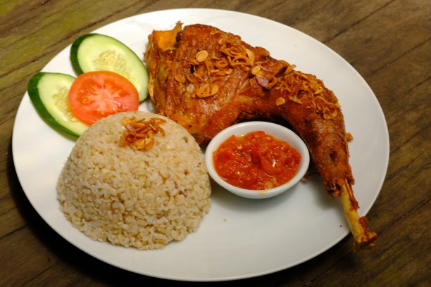 Ayam Kampung Goreng是一种印尼炸鸡，配上糙米、新鲜蔬菜(如西红柿和黄瓜)和辣椒酱。在一张棕色的木桌上。印尼的食物。图片下载