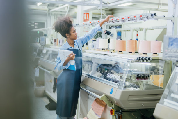 美国女设计师的照片。她穿着制服，拿着订单单，站在缝纫工业工厂里控制工作，检查各种编织设备。图片下载