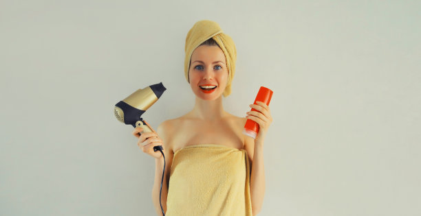 一个快乐微笑的年轻女子的肖像，她在洗澡后拿着一瓶洗发水，用裹好的浴巾擦干湿头发图片下载