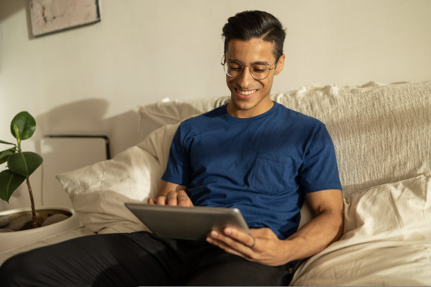 有魅力的西班牙裔男子戴着眼镜，穿着蓝色衬衫，微笑着坐在公寓的沙发上使用平板电脑图片下载