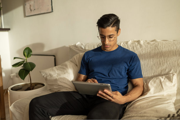 有魅力的西班牙裔男子戴着眼镜，穿着蓝色衬衫，坐在公寓的沙发上使用平板电脑图片下载