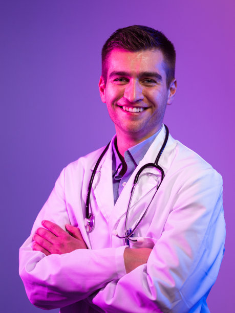 穿白大褂的英雄画像。在蓝色和粉色的背景下，医院里带着听诊器的年轻医生微笑着站着。冠状病毒covid-19危险警报图片下载