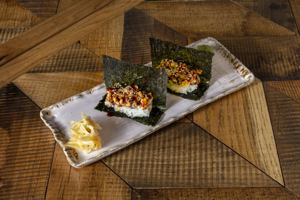 用手卷蘸着照烧酱的鳗鱼，撒上芝麻，放在海苔上。手卷放在一个长方形的陶瓷盘子里，旁边放着芥末和腌姜。盘子放在有图案的木桌上。图片下载