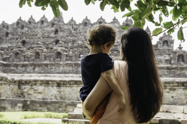 一名亚洲中国妇女抱着她的儿子走在草地上，背景是婆罗浮屠寺图片下载