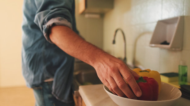 在公寓的厨房里，穿着牛仔衬衫的男人洗蔬菜的手的特写，蔬菜在前景图片下载
