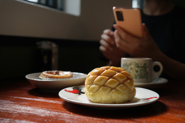 一位年轻的女游客用她的智能手机拍下了美味的食物，港式马球包和蛋挞，还有一杯牛奶咖啡放在木桌上。图片下载