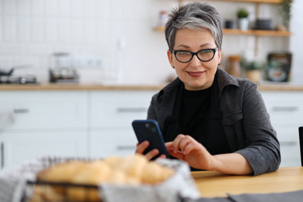 一位面带微笑、手里拿着手机的年长女性坐在餐桌旁，背景是一间家庭厨房。图片下载