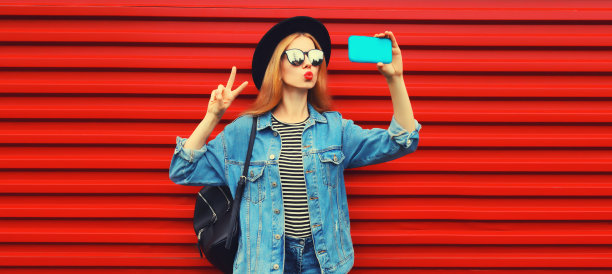 美丽的年轻女子的照片，她吹着嘴唇，用智能手机自拍，穿着牛仔夹克，戴着红色背景的黑色圆帽图片下载