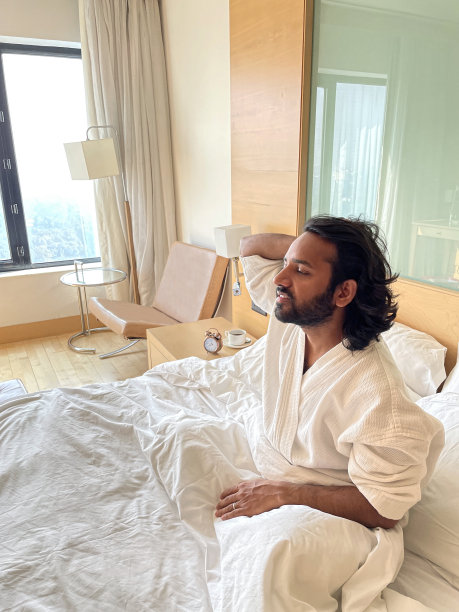 特写照片:印度男子将手臂伸过头顶醒来，穿着白色毛巾浴衣坐在床上，酒店双人床的木质和玻璃床头板，一堆枕头，床头柜上有闹钟的咖啡杯和茶托，重点放在前景上图片下载