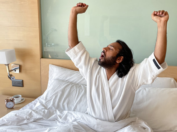 特写照片:印度男子将手臂伸过头顶，打着哈欠，穿着白色毛巾浴衣坐在床上，酒店双人床的木质和玻璃床头板，一堆枕头，床头柜上有闹钟的咖啡杯和茶托，重点放在前景上图片下载