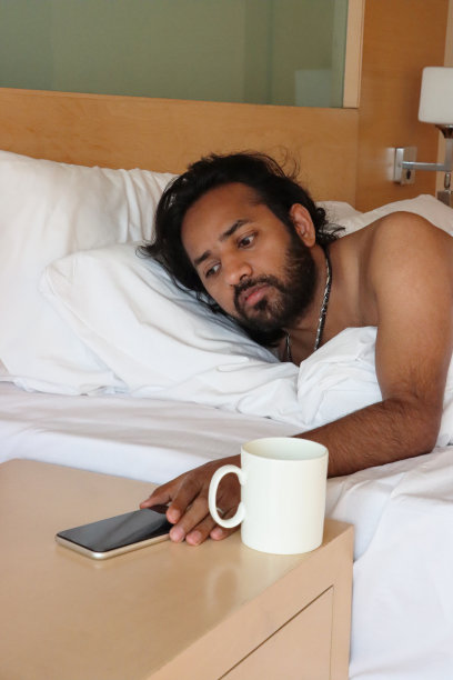 印度男子从羽绒被下穿过酒店双人床，拿着咖啡杯关掉床头柜上的智能手机闹钟的特写照片，重点放在前景图片下载